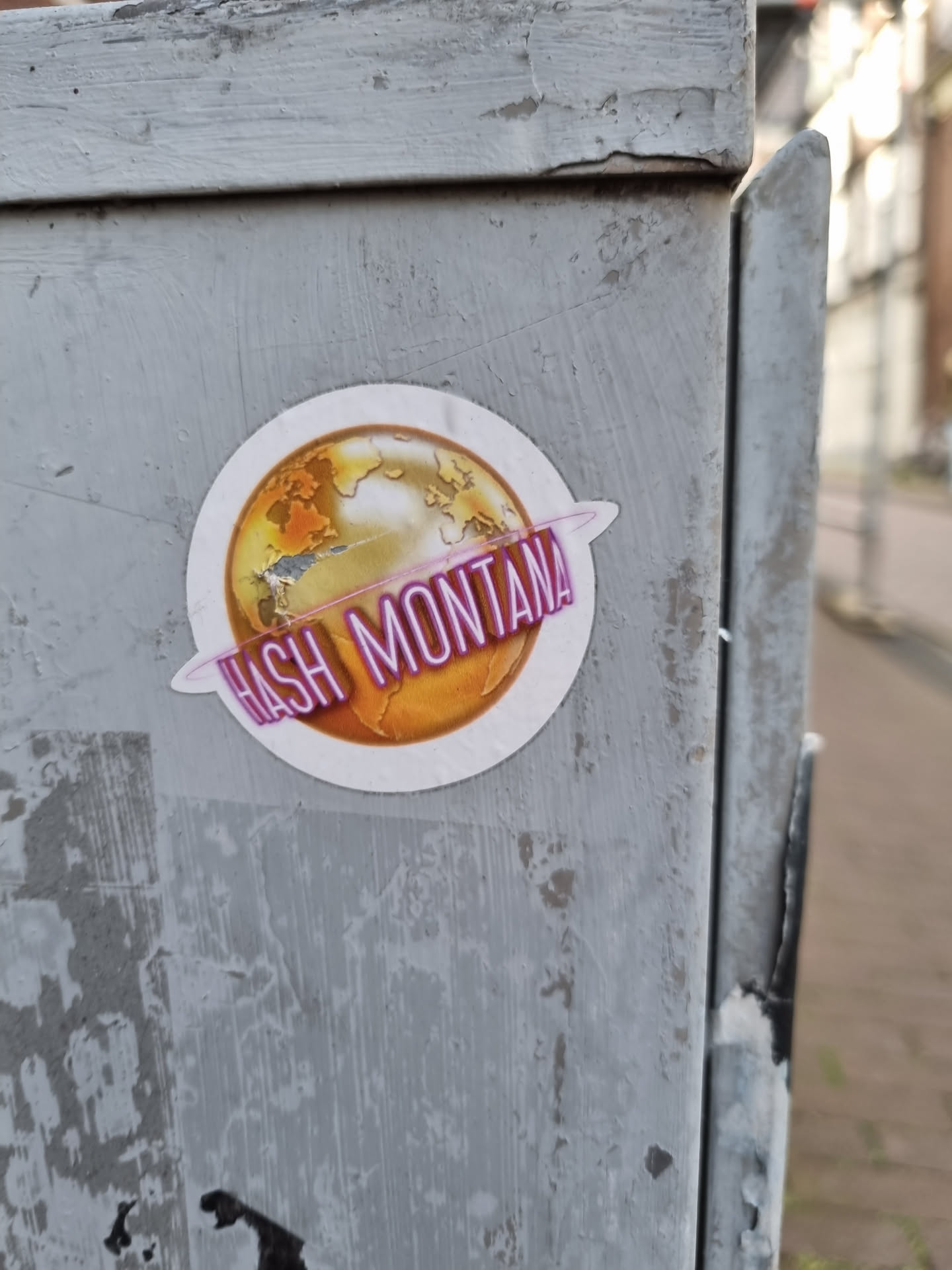 Round sticker with a golden globe and the text Hash Montana written in a purple neon-like style. The globe is stylized and has continents outlined in darker gold.