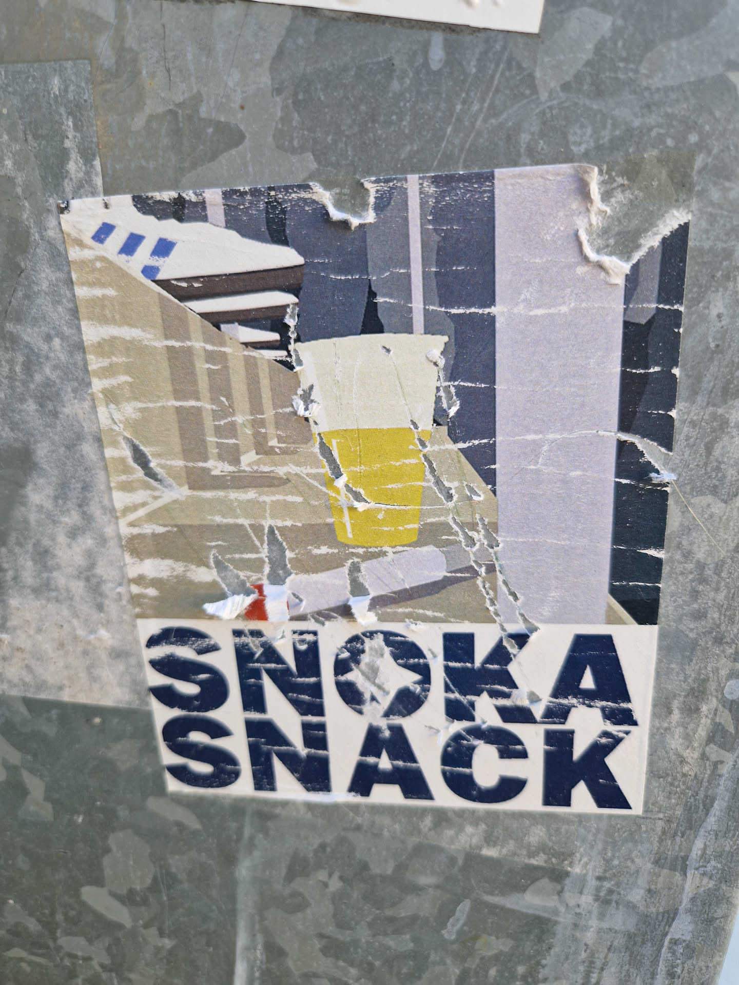 A torn sticker with an illustration of a drink and the text Snoka Snack. The sticker shows signs of wear and tear, with parts of the image missing.