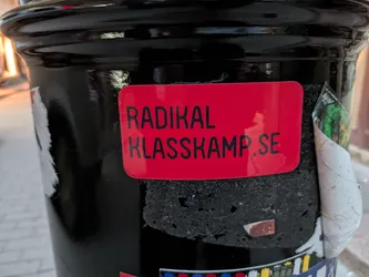 The sticker is a vibrant red rectangle with rounded corners. It features black, sans-serif text that reads "RADIKAL KLASSKAMP.SE" in two lines. The sticker appears to be slightly peeling at the edges, revealing the textured black surface it is adhered to. A portion of another, unidentifiable sticker or graphic is visible to the right, with green and yellow elements and a curled edge.