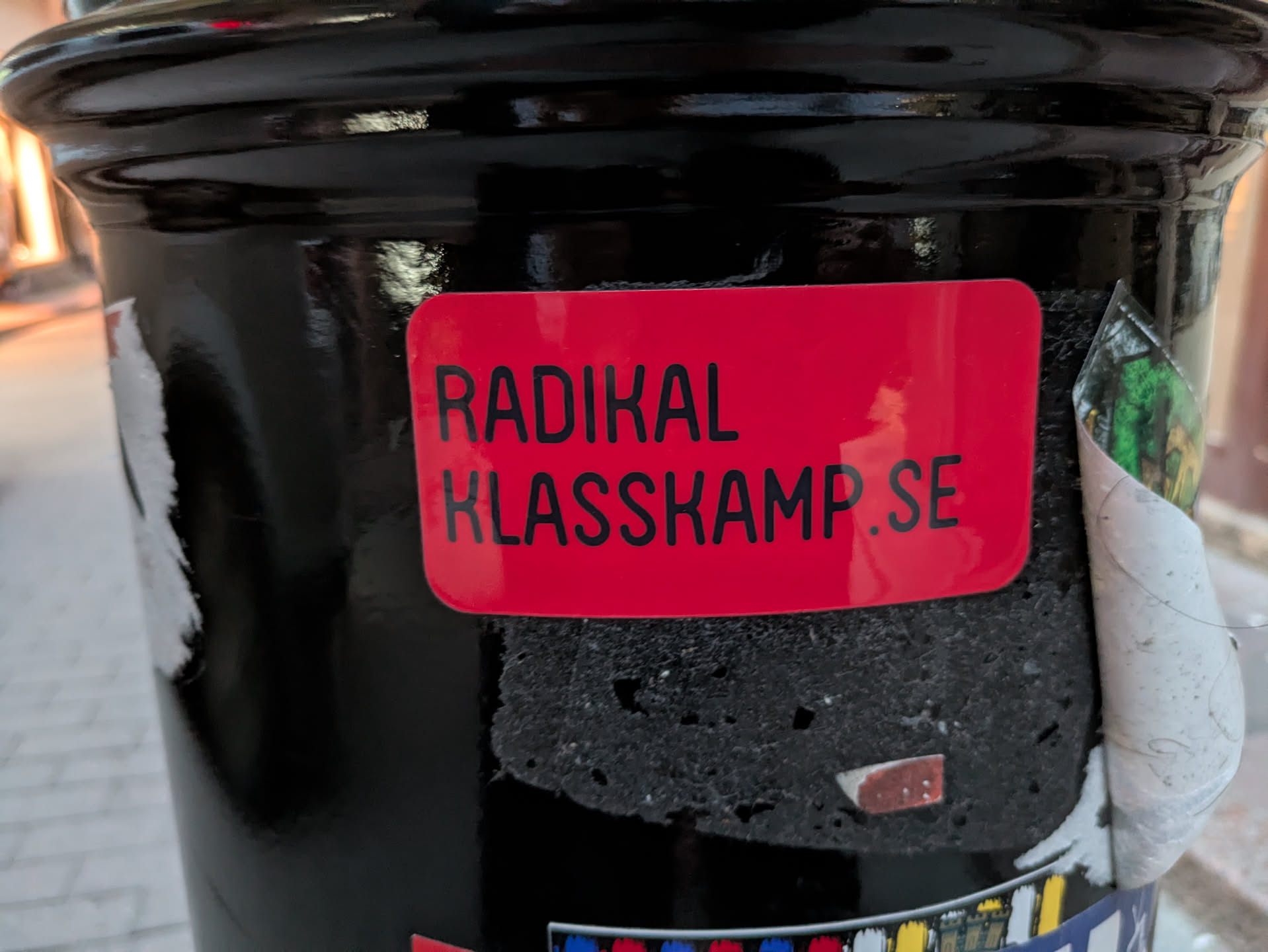 The sticker is a vibrant red rectangle with rounded corners. It features black, sans-serif text that reads "RADIKAL KLASSKAMP.SE" in two lines. The sticker appears to be slightly peeling at the edges, revealing the textured black surface it is adhered to. A portion of another, unidentifiable sticker or graphic is visible to the right, with green and yellow elements and a curled edge.