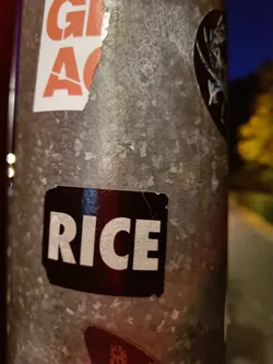 A black sticker with the word "RICE" in white capital letters is attached to a metallic surface.  The sticker shows some signs of wear and tear.  The background features other stickers.