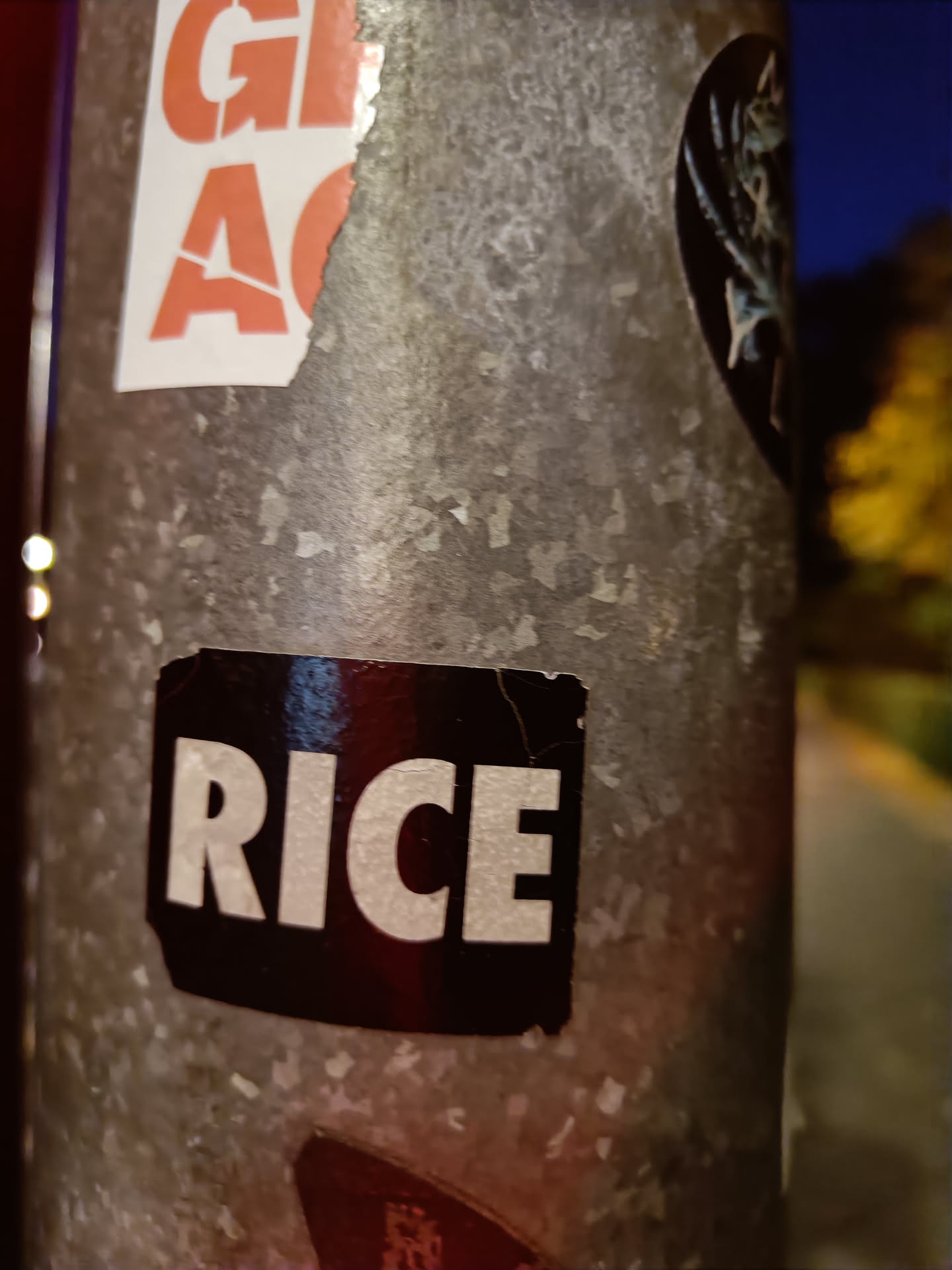 A black sticker with the word "RICE" in white capital letters is attached to a metallic surface.  The sticker shows some signs of wear and tear.  The background features other stickers.