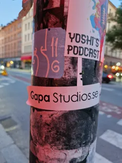 A sticker on a pole advertising Gapa Studios with a website address. Another sticker nearby mentions Yoshi's Podcast.