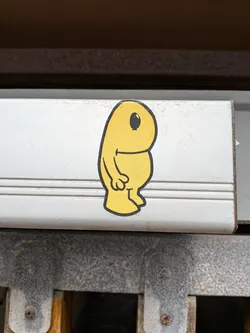 The sticker features a stylized, plump yellow character with a rounded head and body. It has a single, large black eye with a white highlight, positioned on the left side of its head. A simple, curved black line suggests a mouth or smile. The character has two small, stubby arms with three-fingered hands, and two small feet. The outline of the character is a thick black line. The sticker is placed on a textured, light-colored surface.
