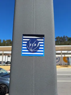 A rectangular sticker with a dark blue border, affixed to a textured grey pole. The background features alternating horizontal blue and white stripes. Superimposed on the blue stripes, the text 'UM OITO NOVE TRÊS' is subtly repeated in a lighter blue. At the center, a large dark blue circle contains a white outline of a stylized ball, resembling a hybrid of a soccer and rugby ball. Inside this ball, the letters 'FCP' are prominently displayed in white capital letters.
