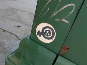 A circular sticker adheres to a weathered, dark green metal surface, likely a utility box or cabinet. The sticker has a white background with a prominent black graphic. This graphic ingeniously combines the traditional female gender symbol (Venus symbol) with a clenched, raised fist integrated into the loop of the symbol, symbolizing female empowerment, solidarity, and resistance. The sticker shows slight signs of wear, including minor cracks on its surface, indicating exposure to the elements. Above the sticker, there are visible white scratch marks on the green surface, and a rusty screw is located to the right.