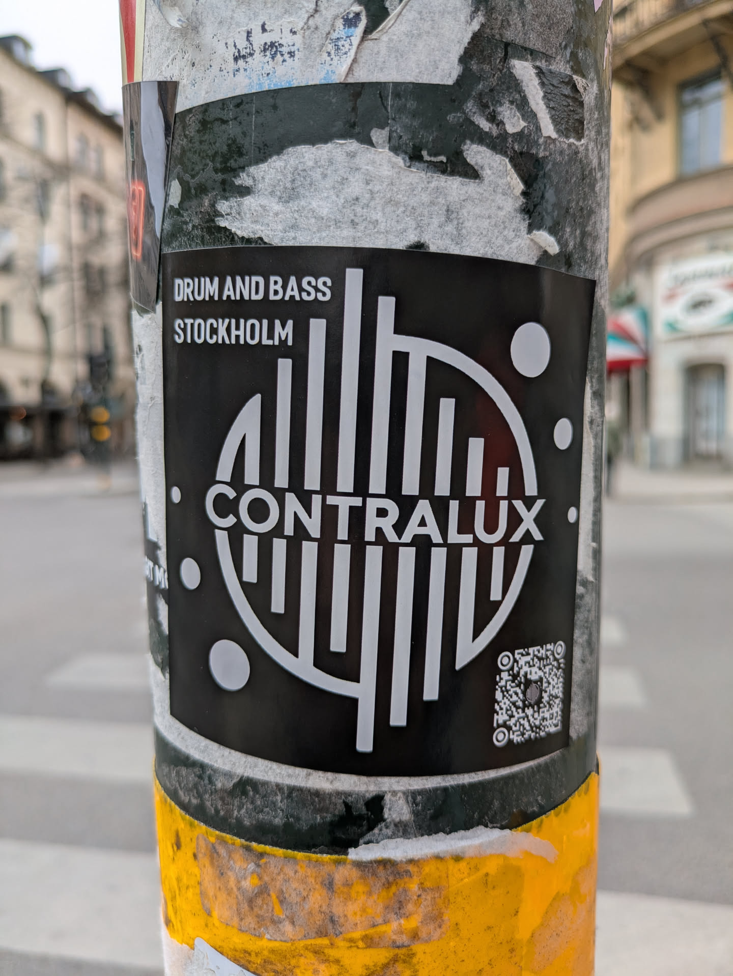A black and white Contralux drum and bass sticker is affixed to a pole in Stockholm. The sticker features the band's name and logo, as well as a QR code.