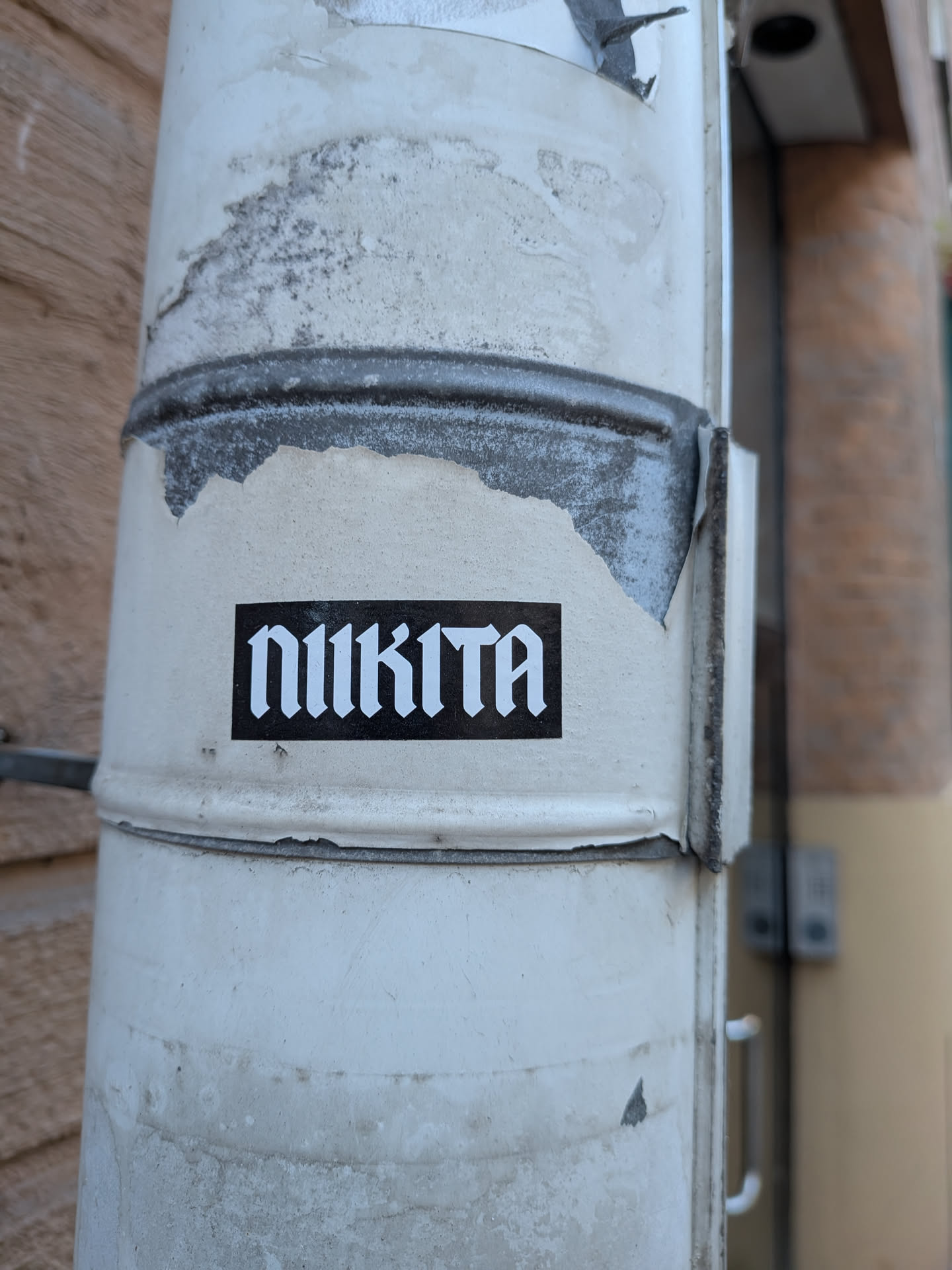 A small horizontal rectangular sticker featuring a solid black background with the word 'NIKITA' written in a bold, stylized white gothic or blackletter font. It is stuck to a worn, peeling white painted downspout.