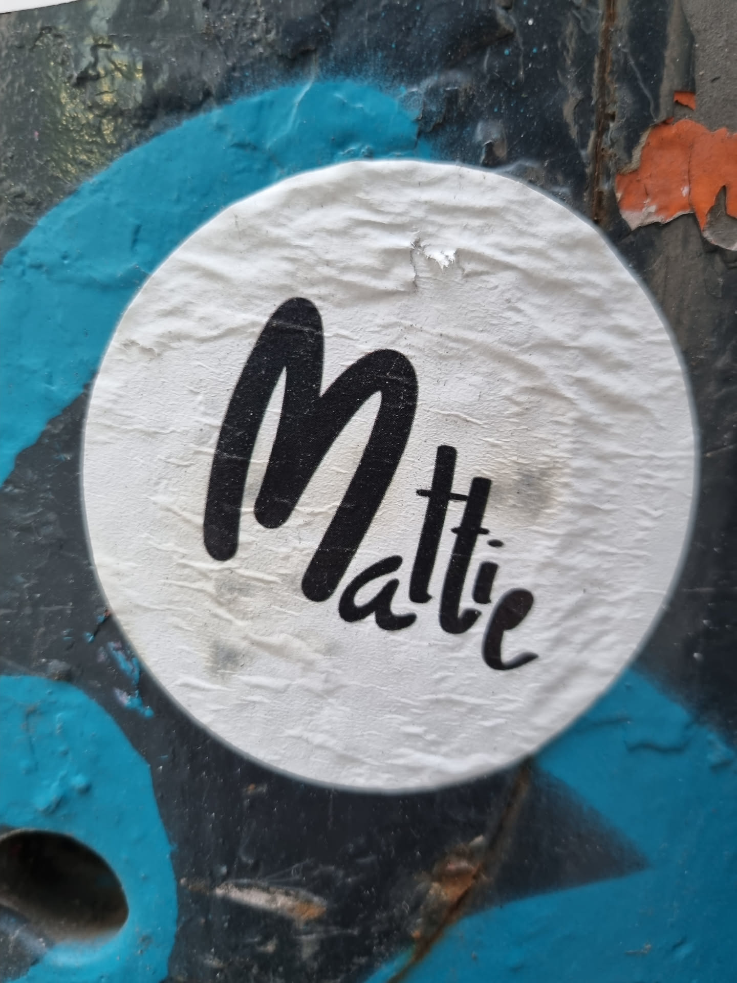 A round white sticker with the name Mattie written in a stylized black script. The sticker is affixed to a surface with teal and black graffiti.