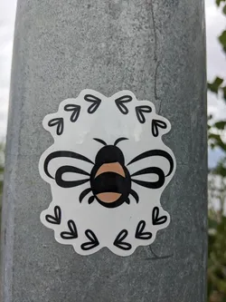 A cute bee sticker with a floral frame. The bee is black and peach in color, and the frame is made of small black hearts and arrows.