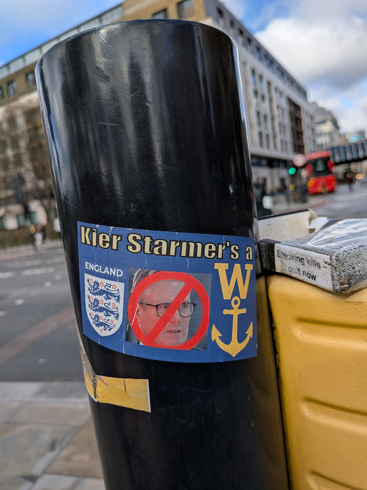 The sticker is rectangular with a blue background. At the top, in yellow capital letters, it reads "Kier Starmer's a W". Below this, on the left, is the word "ENGLAND" above the white and red crest of the England national football team, featuring three lions. In the center, there's a headshot of Keir Starmer, a man with glasses, overlaid with a prominent red circle and a diagonal line, indicating a 'no' or 'forbidden' symbol. To the right of Starmer's image, there's a large yellow capital 'W' followed by a yellow anchor symbol. The sticker shows signs of wear, with some creases and slight peeling, particularly at the bottom edge, suggesting it has been exposed to the elements.