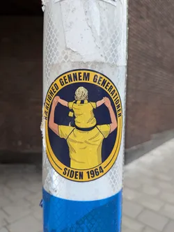 Street sticker An oval sticker featuring a yellow and dark blue color scheme, associated with the Danish football club Brøndby IF. The illustration shows a father viewed from behind carrying a small child on his shoulders, both wearing matching yellow football shirts with dark blue collars. Around the top arch of the yellow border, the Danish text reads 'KÆRLIGHED GENNEM GENERATIONER', while the bottom arch reads 'SIDEN 1964'.