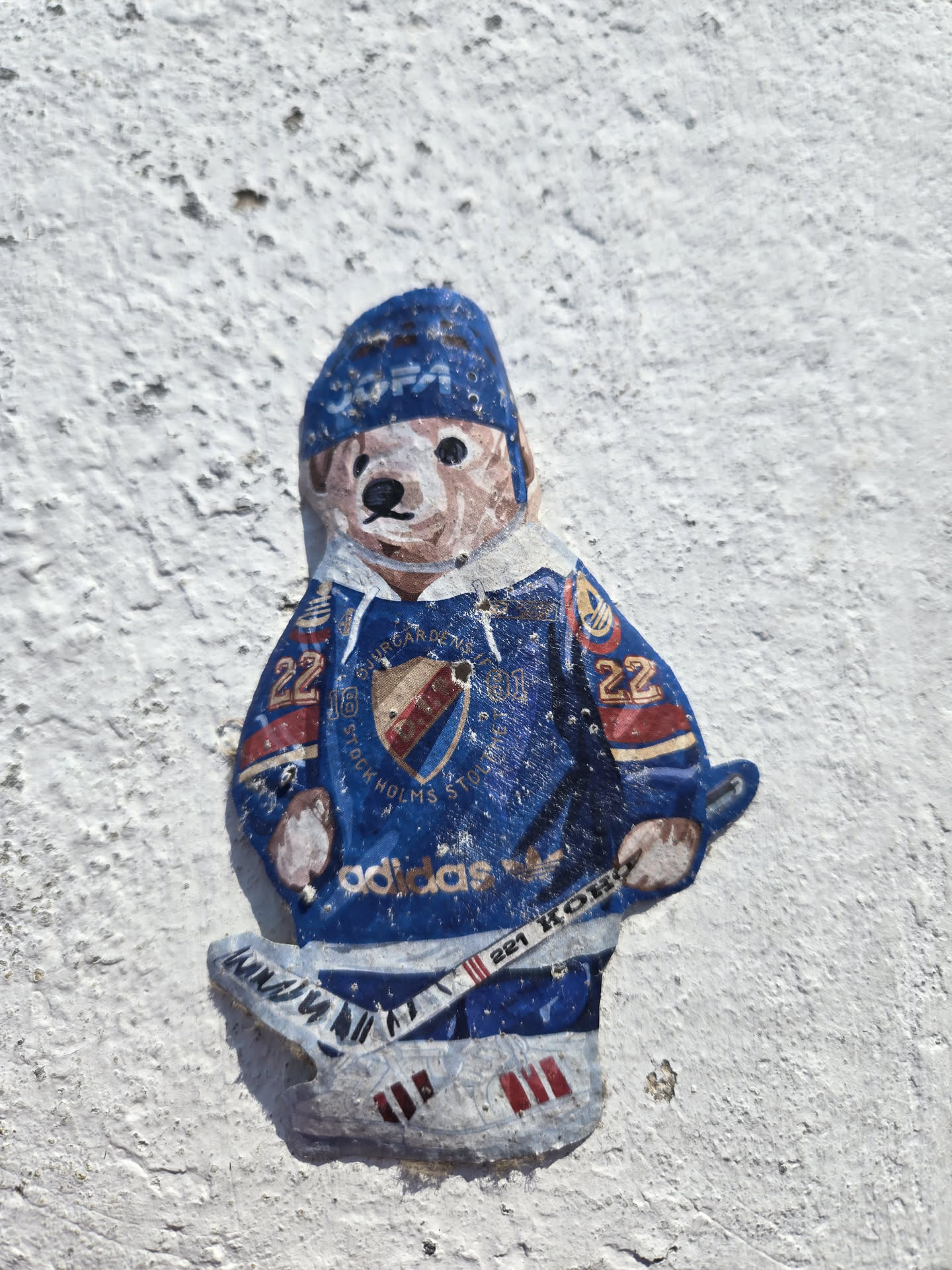 The sticker depicts a cartoonish bear wearing a blue hockey uniform. The bear has a light brown face with black eyes and nose, and a small smile. It wears a blue beanie with white lettering that is partially obscured. Its jersey is predominantly blue with red and white accents, featuring the team logo on the chest, which appears to be a shield with red and gold elements and possibly a crown. The jersey also has the number '22' on the sleeves. The bear is holding a hockey stick, and its feet are depicted in hockey skates with red Adidas stripes on the sides. The overall style is somewhat naive and hand-drawn, with visible texture suggesting it's a physical sticker applied to a rough, white surface.
