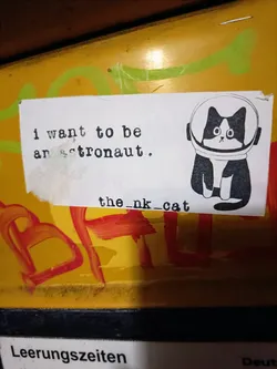 A sticker featuring a black and white cat wearing an astronaut helmet. The text 'I want to be an astronaut' is written above the cat, and 'the_nk_cat' is written below it.