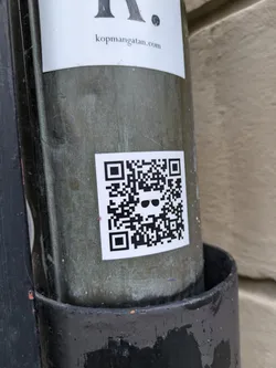 A small, square sticker featuring a QR code with a sunglasses emoji in the center is affixed to a weathered pole. The sticker is predominantly black and white, with the QR code taking up most of the space. The sunglasses emoji is simple, adding a touch of character to the otherwise functional design. The pole appears to be made of metal, showing signs of age and exposure to the elements.