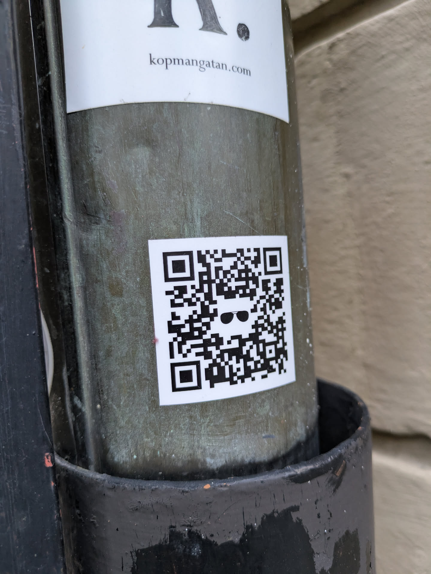 A small, square sticker featuring a QR code with a sunglasses emoji in the center is affixed to a weathered pole. The sticker is predominantly black and white, with the QR code taking up most of the space. The sunglasses emoji is simple, adding a touch of character to the otherwise functional design. The pole appears to be made of metal, showing signs of age and exposure to the elements.