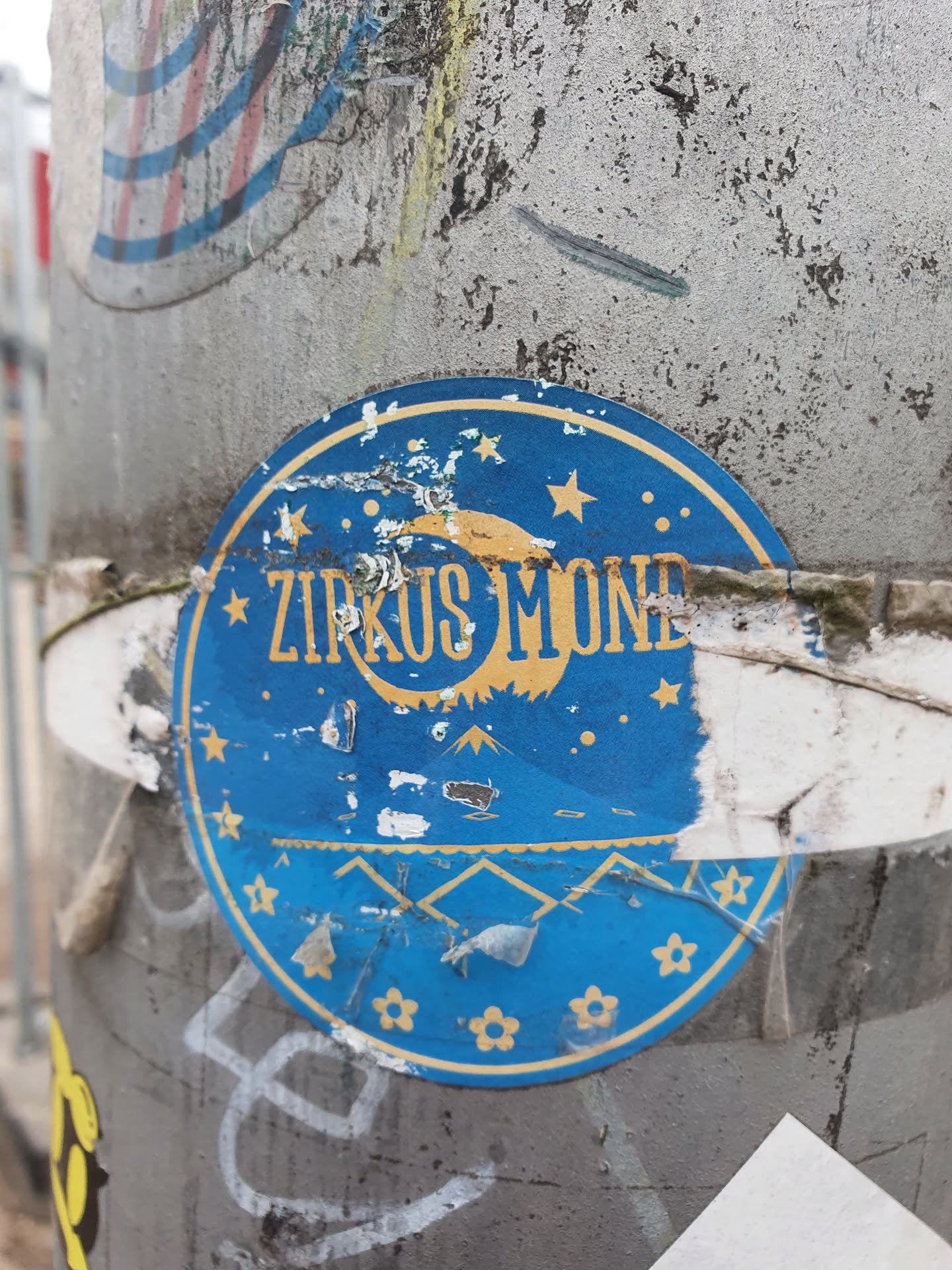 A round blue sticker with the text Zirkus Mond, a yellow moon and stars. It's partially damaged and peeling off a grey pole.