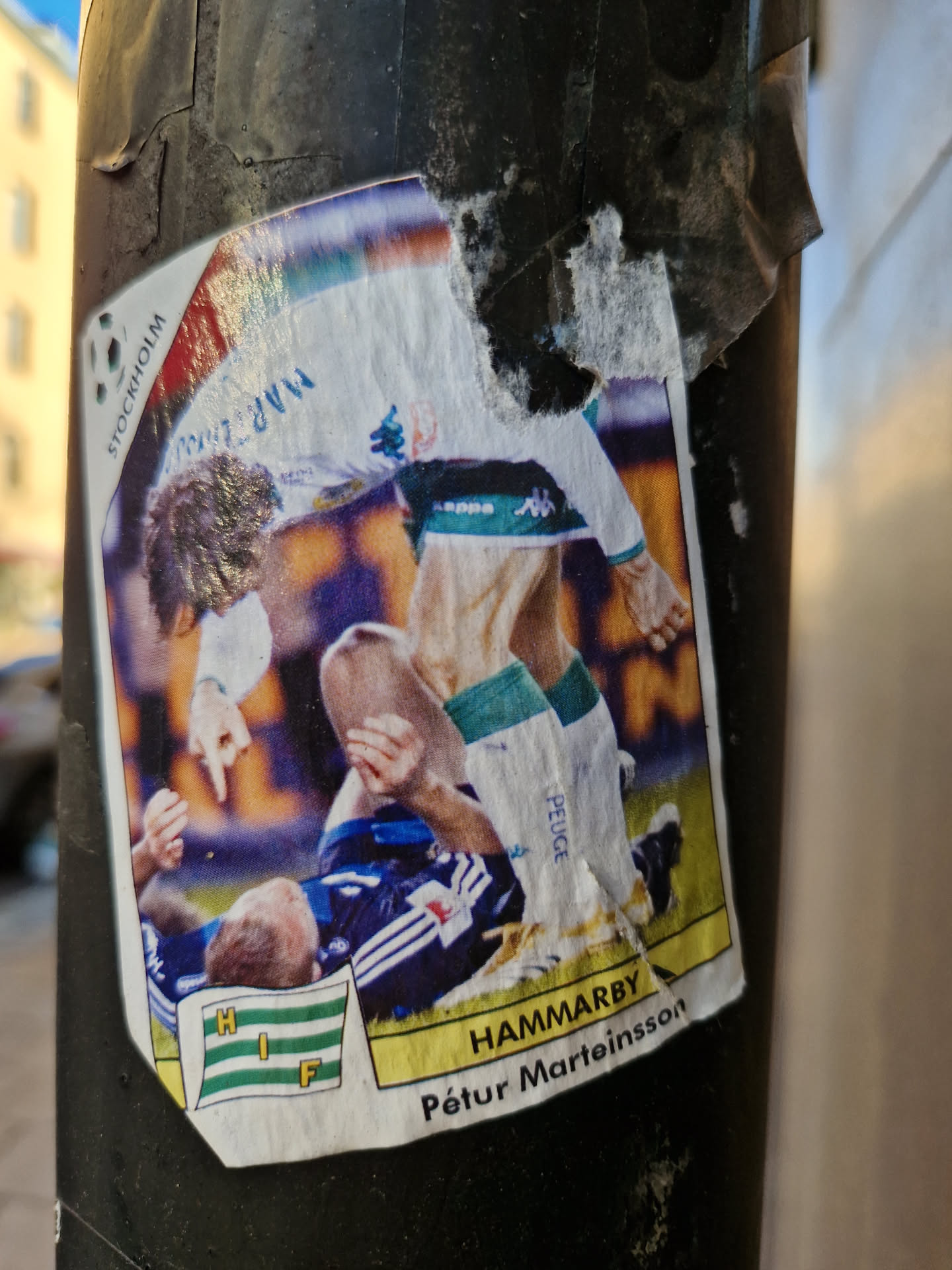 A sticker depicting Hammarby footballer Pétur Marteinsson in action. The sticker shows a dynamic scene from a football match, with the player in a challenging moment.  The sticker is slightly worn and torn around the edges, suggesting it has been affixed outdoors for some time.