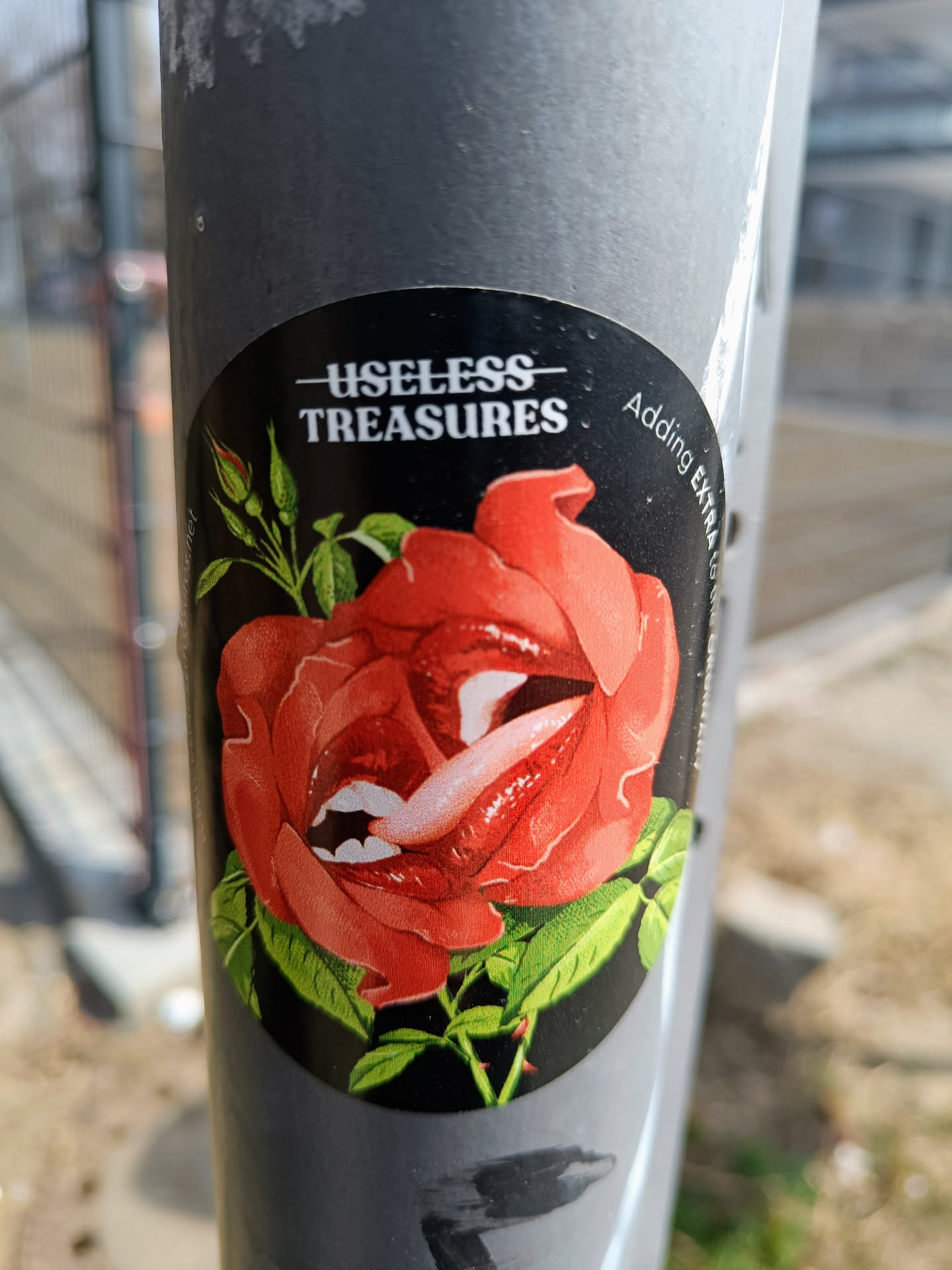 A round sticker featuring a red rose with a tongue sticking out. The text \"Useless Treasures\" is written at the top. \"Adding extra\" is written on the right side.