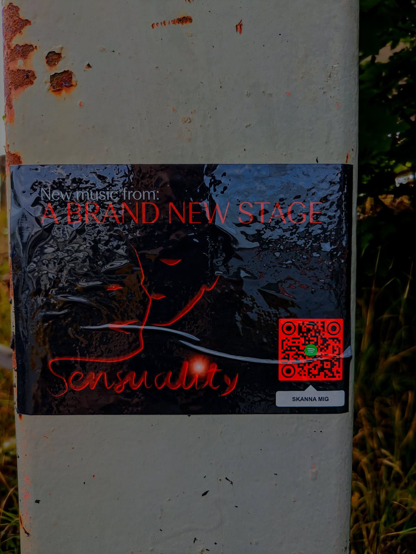 A dark, glossy sticker is adhered to a weathered, rust-stained white pole outdoors. The sticker features a striking design with red text and graphics against a black background that appears to have an undulating, liquid-like texture. At the top, 'New music from:' is written in white, followed by 'A BRAND NEW STAGE' in bold, red uppercase letters. Below this, an abstract outline of a profile or face, rendered in red, merges with the textured background. Towards the bottom left, the word 'Sensuality' is written in a flowing, stylized red script, accompanied by a subtle red glow. On the right side, a prominent red QR code with a green Spotify logo at its center is visible. Beneath the QR code, a small grey rectangular button contains the text 'SKANNA MIG' in white.