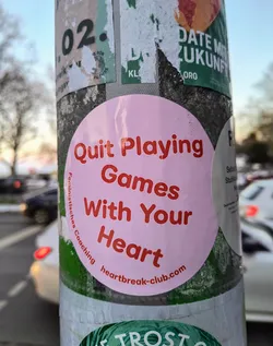 A round, pink sticker with red text is adhered to a pole. The text reads "Quit Playing Games With Your Heart" and "Feministisches Coaching heartbreak-club.com".
