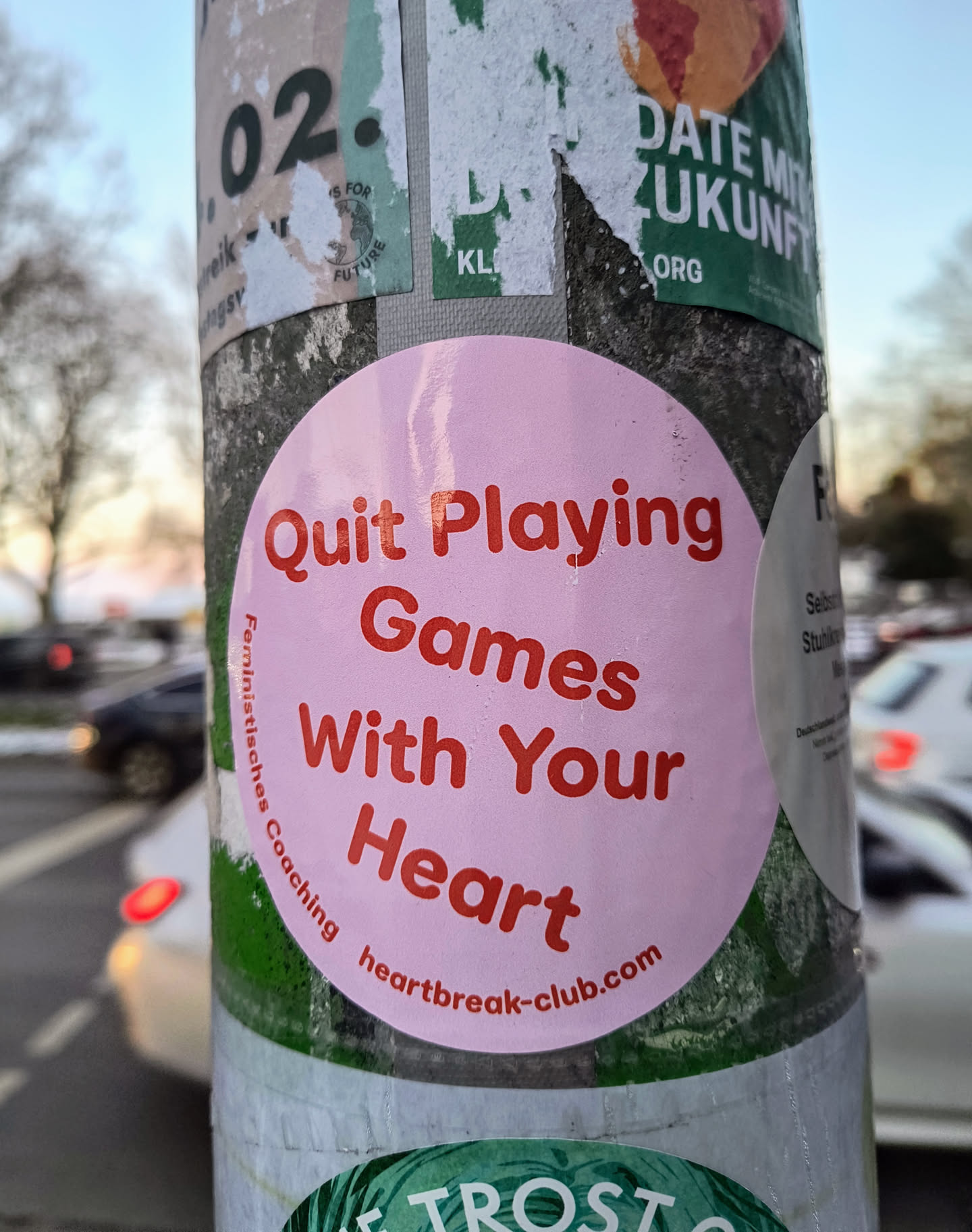 A round, pink sticker with red text is adhered to a pole. The text reads "Quit Playing Games With Your Heart" and "Feministisches Coaching heartbreak-club.com".