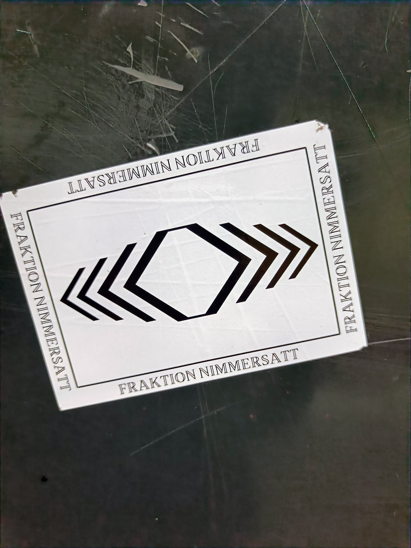 A white square sticker with black graphic design and text. The text 'Fraktion Nimmersatt' is repeated on all four sides. The central design is a geometric pattern of arrows and hexagons.