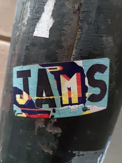 A colorful sticker with the word JAMS in a stylized design. The sticker has a torn and weathered look, adding to its urban aesthetic. The background of the sticker is teal, and the letters are in various colors, primarily black, yellow, and red. There is some small illegible text at the bottom. The sticker is affixed to a dark gray, rough textured surface, possibly a pole or wall. 