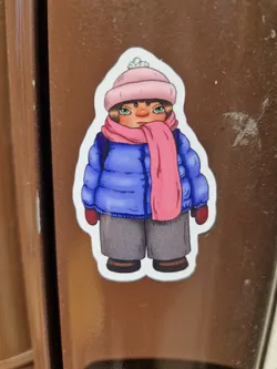 A sticker featuring a cartoon illustration of a girl wearing a pink hat, blue jacket, and pink scarf. The style is simplistic and charming. 