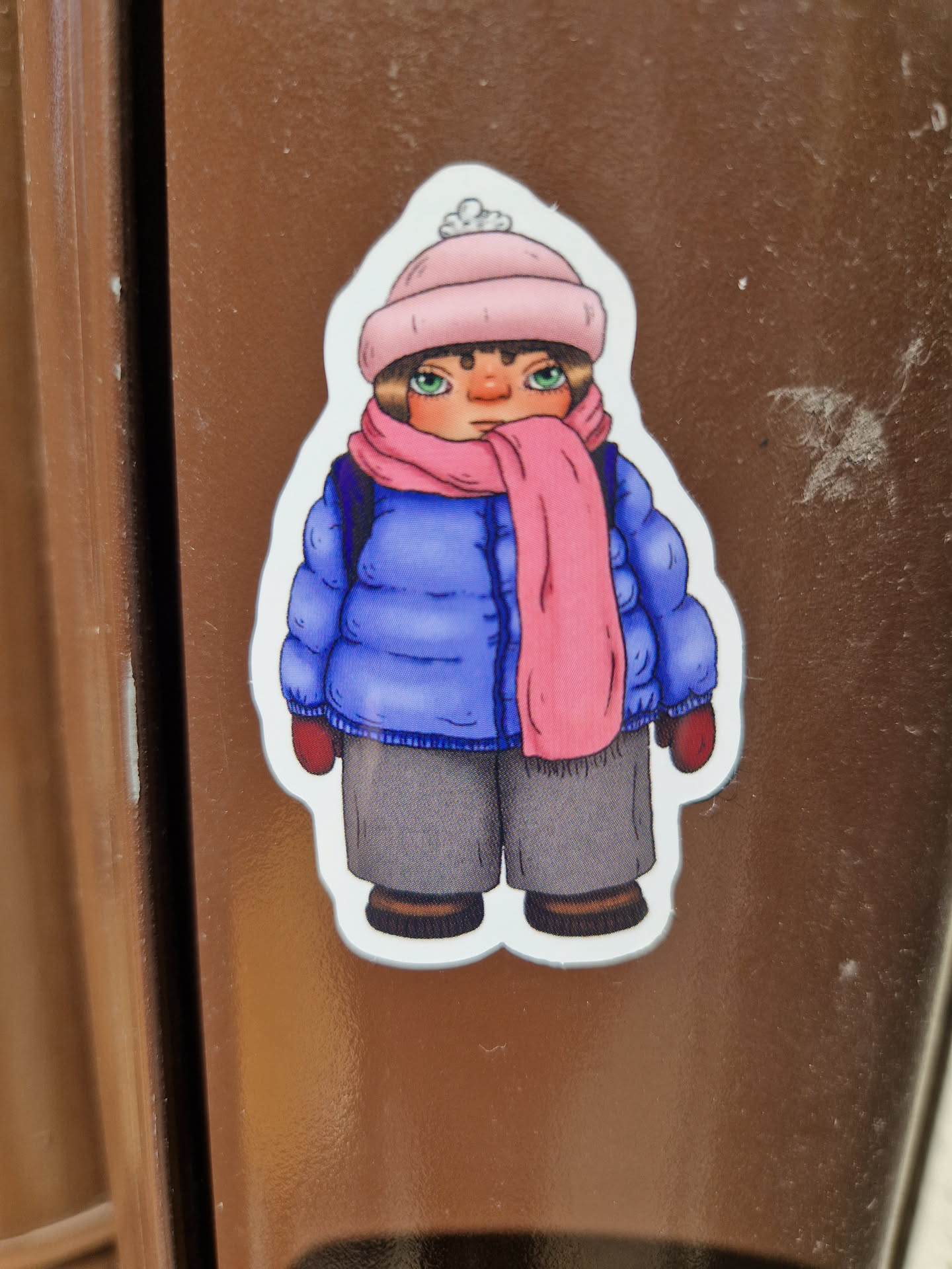 A sticker featuring a cartoon illustration of a girl wearing a pink hat, blue jacket, and pink scarf. The style is simplistic and charming. 