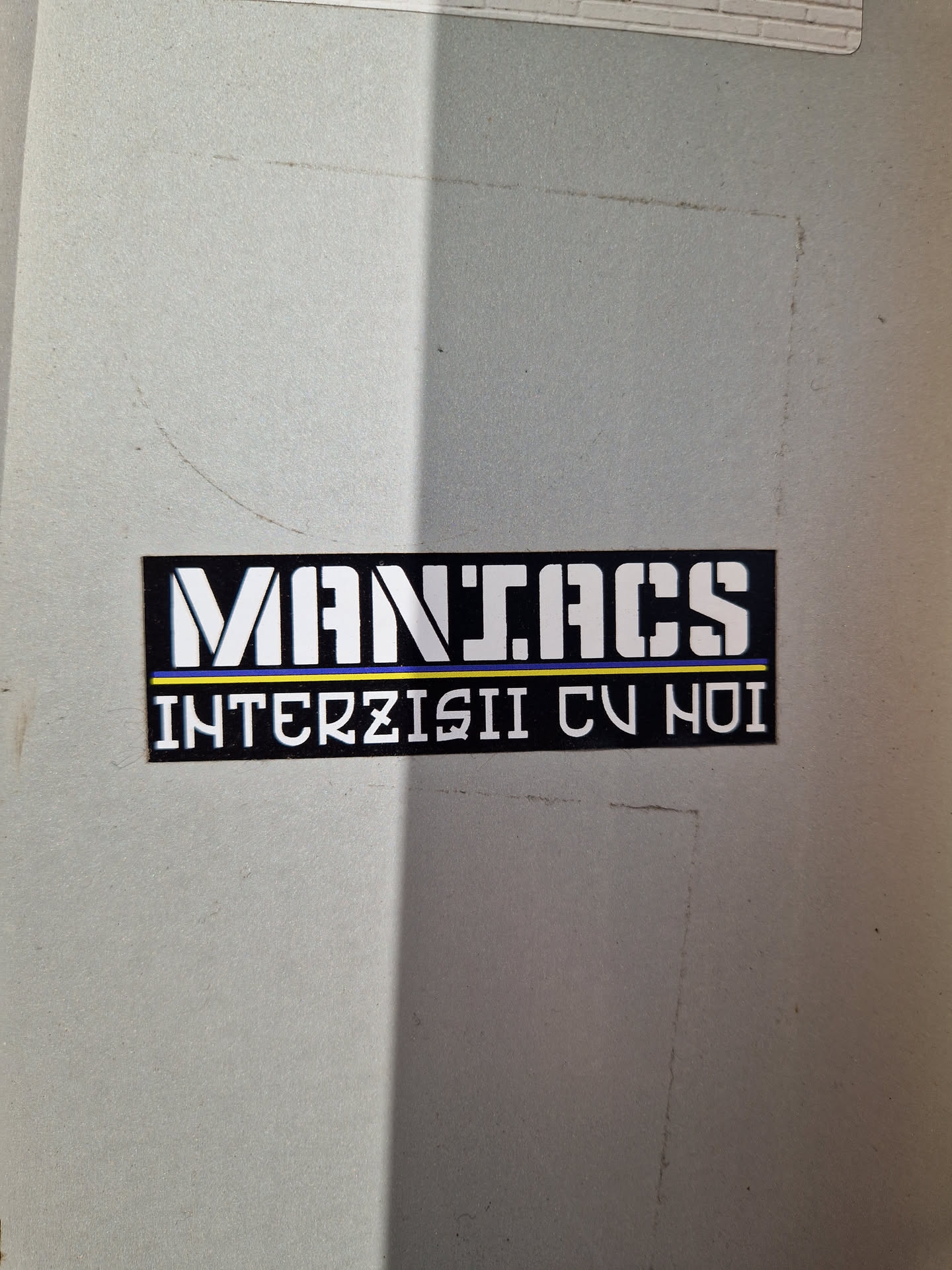 A black and white sticker with the words "MANIACS" in a large, bold font on top, and "INTERZISII CU HOI" in a smaller font underneath. The sticker has a simple design and appears to be placed on a light gray surface.