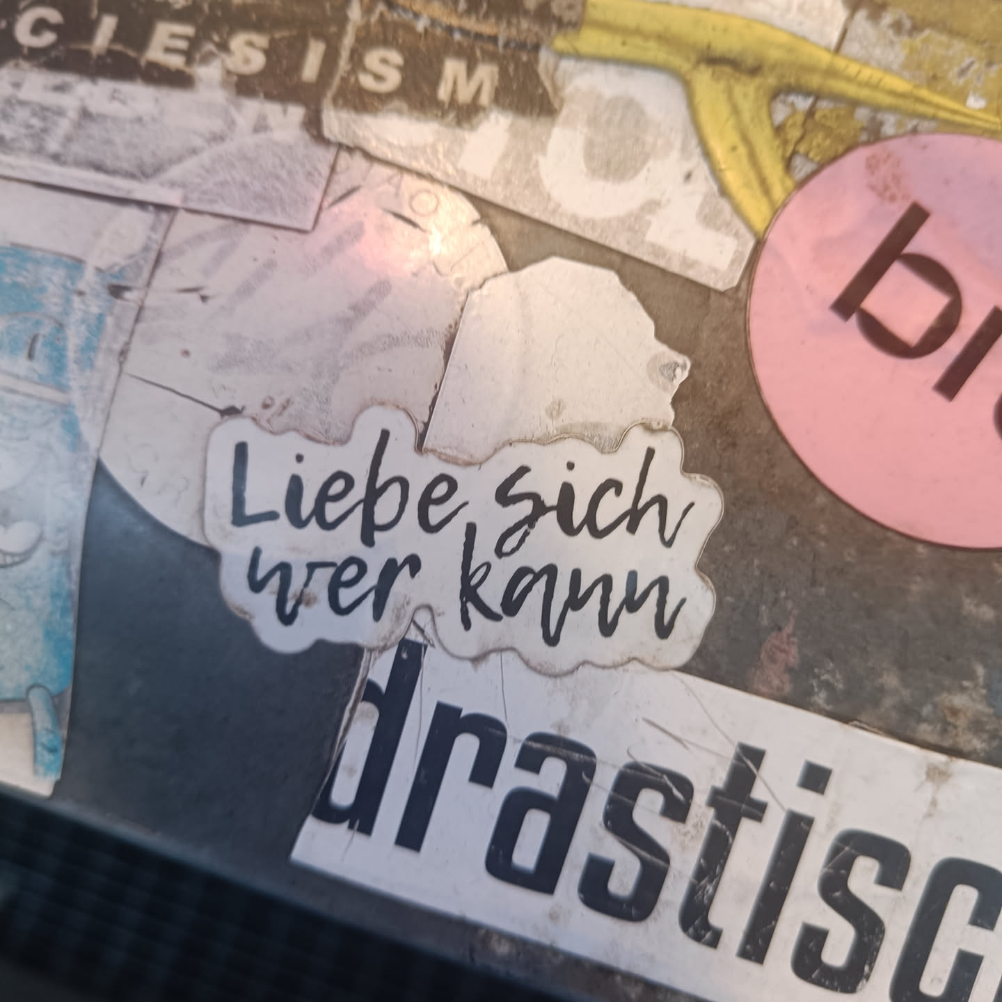 A white sticker with black text on it that says "Liebe sich wer kann" which translates to "Love yourself if you can" in German. The sticker is placed on a surface with other stickers.