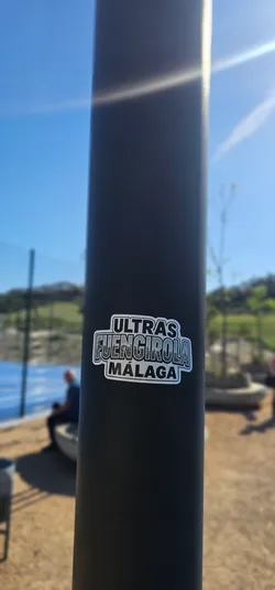 The sticker is a rectangular design with rounded corners, featuring a monochrome palette of black, white, and shades of gray. The top line prominently displays the word "ULTRAS" in a bold, blocky, outlined font. Below this, the word "FUENGIROLA" is rendered in a larger, stylized font, with the letters filled with a detailed grayscale illustration of a cityscape, depicting buildings and structures. The bottom line reads "MÁLAGA" in a font similar to "ULTRAS", also bold and outlined. The entire sticker is bordered by a clean white outline, making it pop against the dark, textured surface of the pole it's affixed to.
