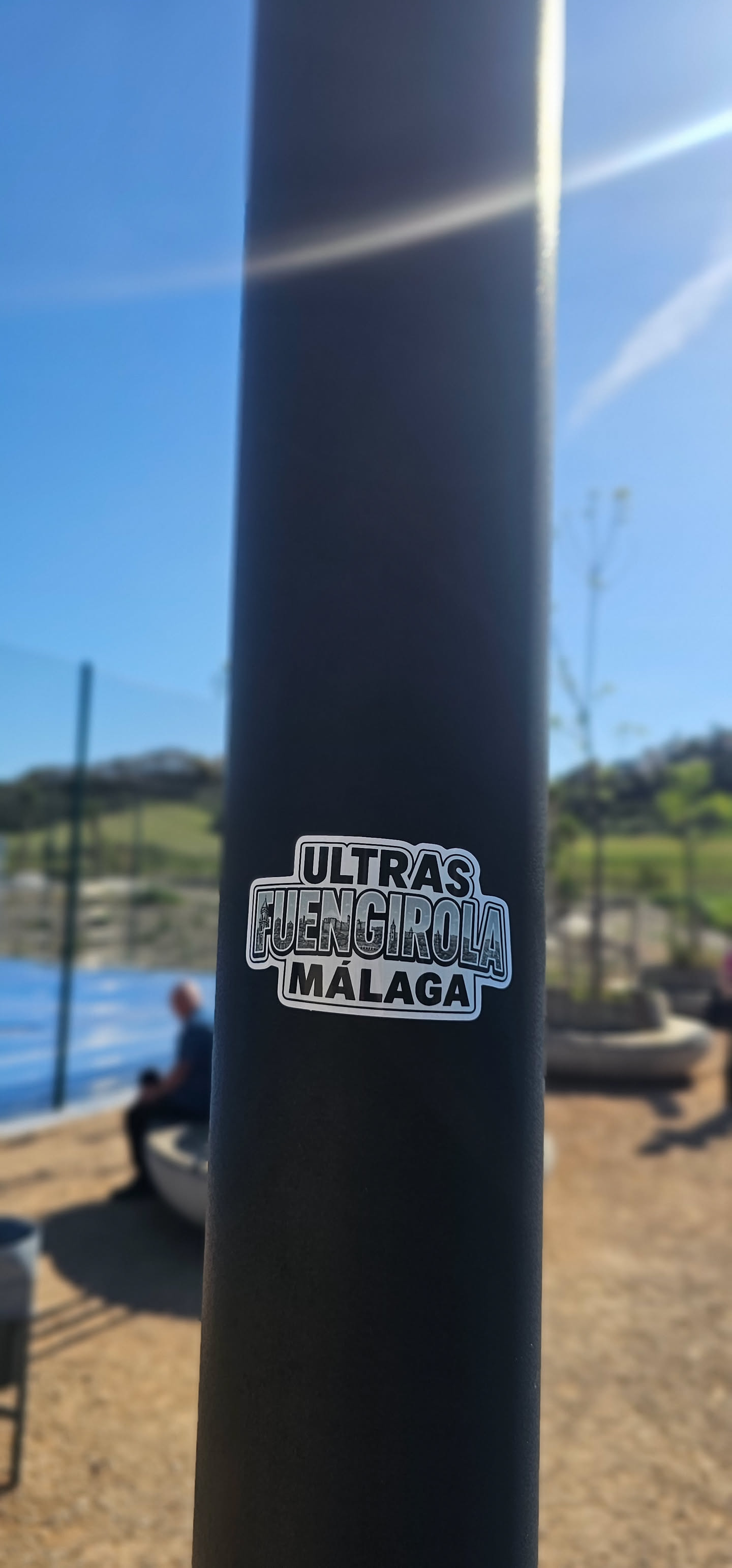 The sticker is a rectangular design with rounded corners, featuring a monochrome palette of black, white, and shades of gray. The top line prominently displays the word "ULTRAS" in a bold, blocky, outlined font. Below this, the word "FUENGIROLA" is rendered in a larger, stylized font, with the letters filled with a detailed grayscale illustration of a cityscape, depicting buildings and structures. The bottom line reads "MÁLAGA" in a font similar to "ULTRAS", also bold and outlined. The entire sticker is bordered by a clean white outline, making it pop against the dark, textured surface of the pole it's affixed to.