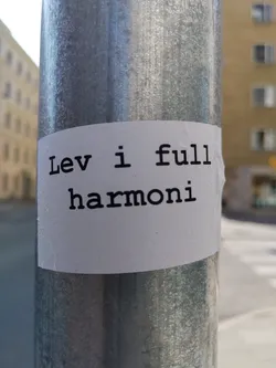 A white sticker on a metal pole with the text "Lev i full harmoni" written in a typewriter-style font.