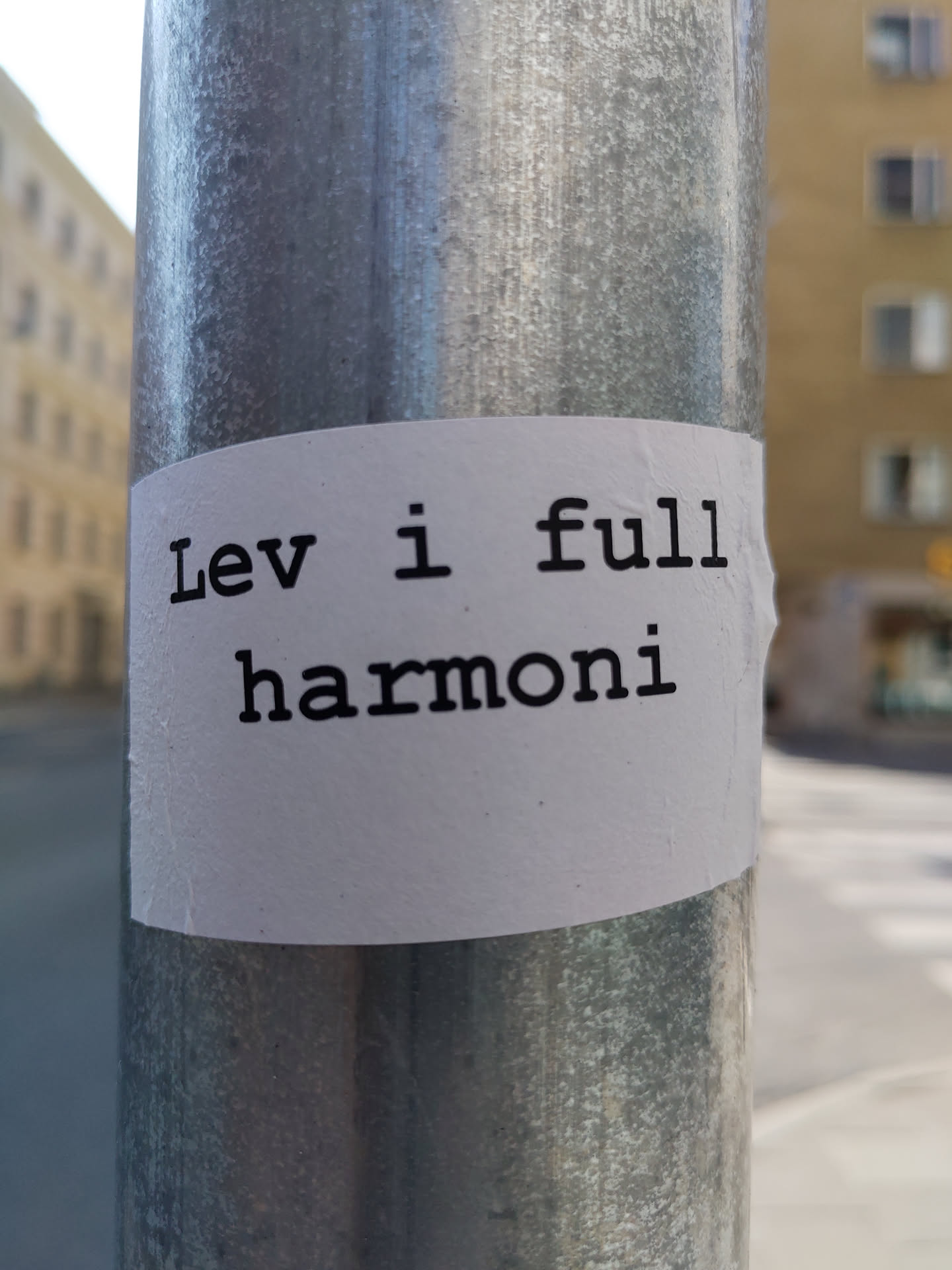 A white sticker on a metal pole with the text "Lev i full harmoni" written in a typewriter-style font.