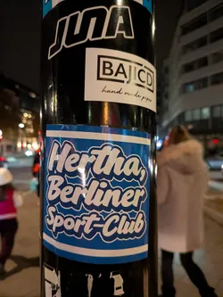 A rectangular sticker adhered to a black lamppost. The sticker features a bold blue background with thin white horizontal bars along the top and bottom edges. Centered on the sticker is the text 'Hertha, Berliner Sport-Club' written in a retro, rounded cursive script. The lettering is white with a thick dark blue outline and shadow effect.