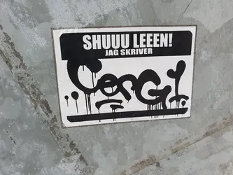 Black and white sticker with graffiti-style lettering. The text at the top says "SHUU LEEEN! JAG SKRIVER" which translates from Swedish to "Shhh read! I write".  The main design is a stylized black graphic resembling a dripping paint effect, possibly a tag or signature.