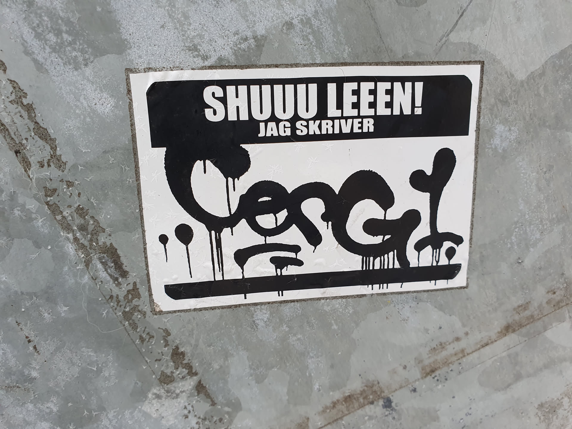 Black and white sticker with graffiti-style lettering. The text at the top says "SHUU LEEEN! JAG SKRIVER" which translates from Swedish to "Shhh read! I write".  The main design is a stylized black graphic resembling a dripping paint effect, possibly a tag or signature.