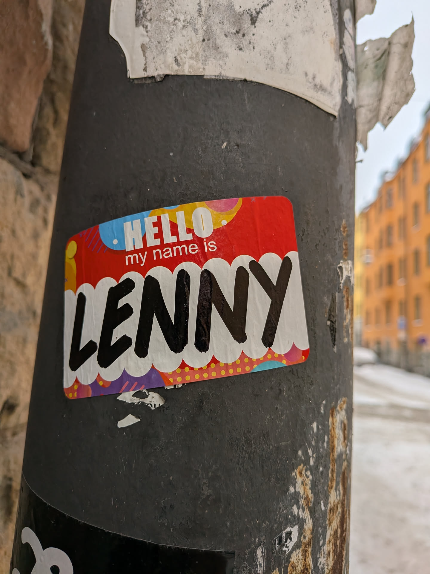 The sticker is a rectangular 'Hello my name is' style tag with rounded corners, adhered to a dark, textured pole. The main background is red. At the top, above 'my name is', there's a colorful, abstract pattern featuring light blue, yellow, pink, and purple shapes with white lines and dots. The word 'HELLO' is prominently displayed in white, bold, sans-serif letters on a light blue background. Below 'HELLO', 'my name is' is written in smaller white sans-serif text on the red background. The name 'LENNY' is written in large, bold, black, bubble-style letters with a distinct white outline, set against a white cloud-like or scalloped shape. Below this white shape, there's another colorful pattern with purple, pink, and light blue elements, including yellow polka dots on a red background. The sticker shows signs of wear, with slight peeling at the edges and some dirt accumulation, indicating it's a genuine street sticker.