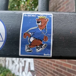 This sticker resembles a postage stamp and showcases a cartoon bear wearing a blue jersey and a crown, walking or running. The text on the sticker indicates '25. Juli 1892' and 'Berlin'.