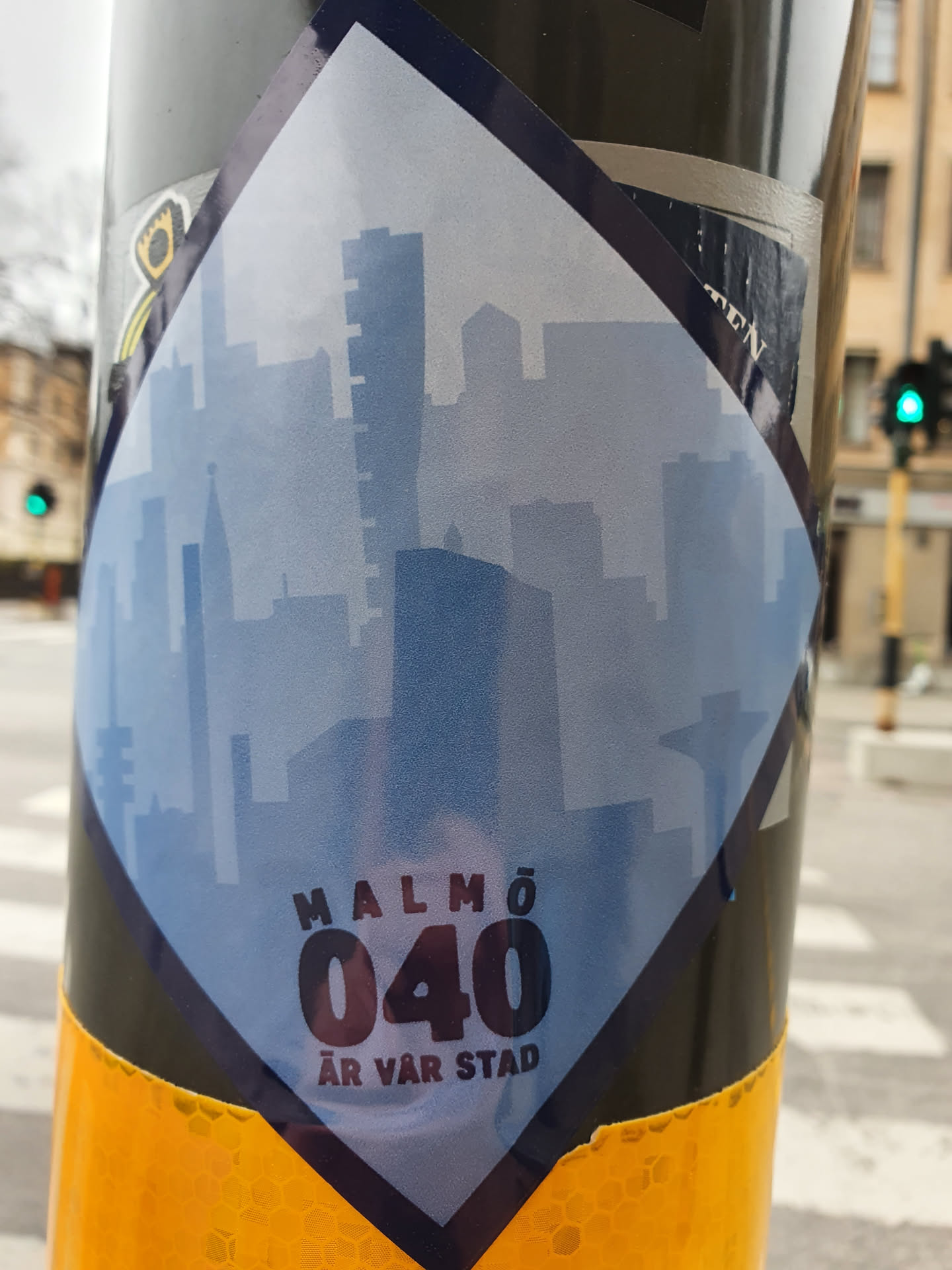 A diamond-shaped sticker depicting a cityscape silhouette of Malmö, Sweden. The text 'MALMÖ 040 ÄR VÅR STAD' is prominently displayed, which translates to 'Malmö 040 is our city'. The sticker has a faded, slightly transparent city skyline design, giving it a subtle, artistic appearance.