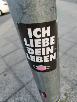A black and white sticker with the text "Ich liebe dein Leben" (I love your life) in German. There is a small pink mask symbol at the bottom.