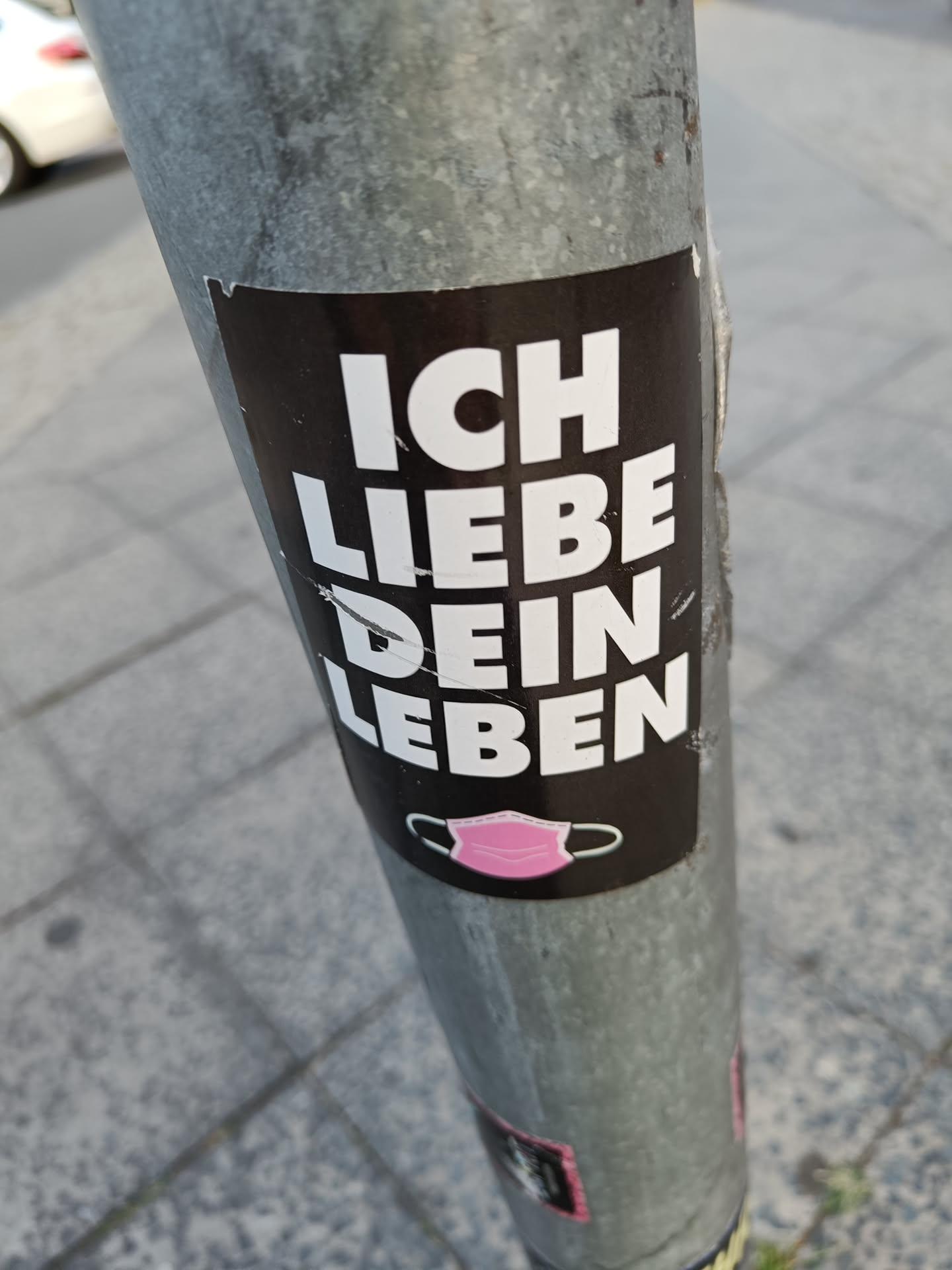 A black and white sticker with the text "Ich liebe dein Leben" (I love your life) in German. There is a small pink mask symbol at the bottom.