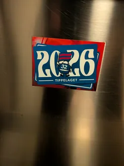 A rectangular sticker with rounded corners set against a brushed metal surface. The sticker features a deep blue background framed by a thin white line. In the center are large stylized white numerals '20' and '26' framing a cartoon character viewed from behind. The stout character wears a blue jersey with '32' printed on the back and waves a horizontally striped red and dark blue flag. Beneath the character, flanked by two horizontal white lines, is the text 'TIPPELAGET' in clean white capital letters.