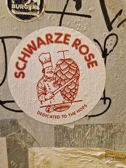 A round sticker featuring a chef preparing a hop cone. The text 'Schwarz Rose' is written in a circular fashion around the illustration, with 'Dedicated to the Hops' at the bottom. The style is reminiscent of vintage brewery advertisements.
