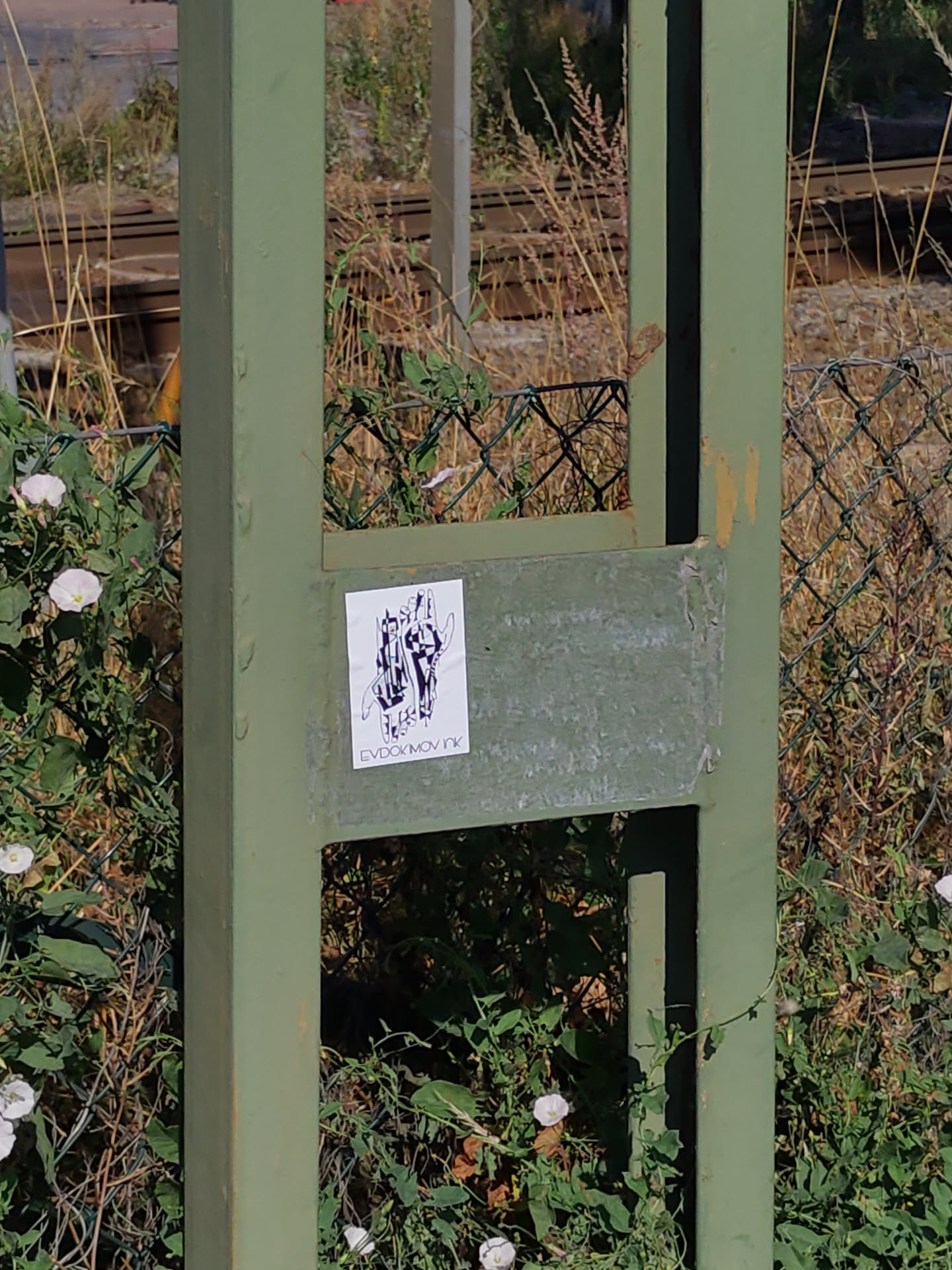 A small, square sticker featuring a black and white illustration of what looks like two figures intertwined. The artist's tag, 'EVDOMVNk', is visible at the bottom. The sticker is affixed to a green metal post near some greenery. 