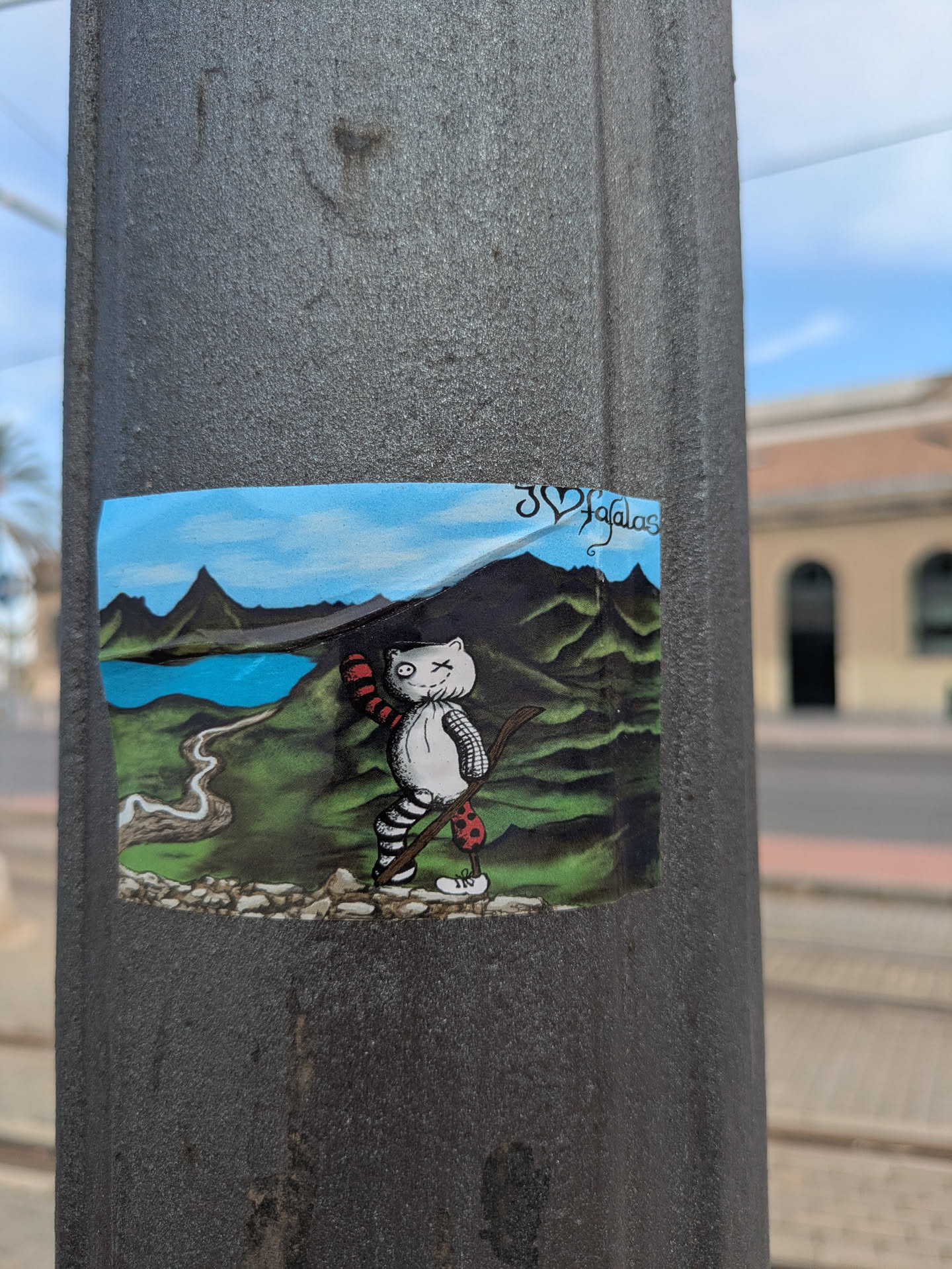 The sticker features a cat-like character with a striped body and red-and-black arm warmer and knee pad, holding a walking stick. It stands in front of a painted landscape with a lake, mountains, and a blue sky. The artist's name is written at the top right corner of the landscape: "3 corações falalas".