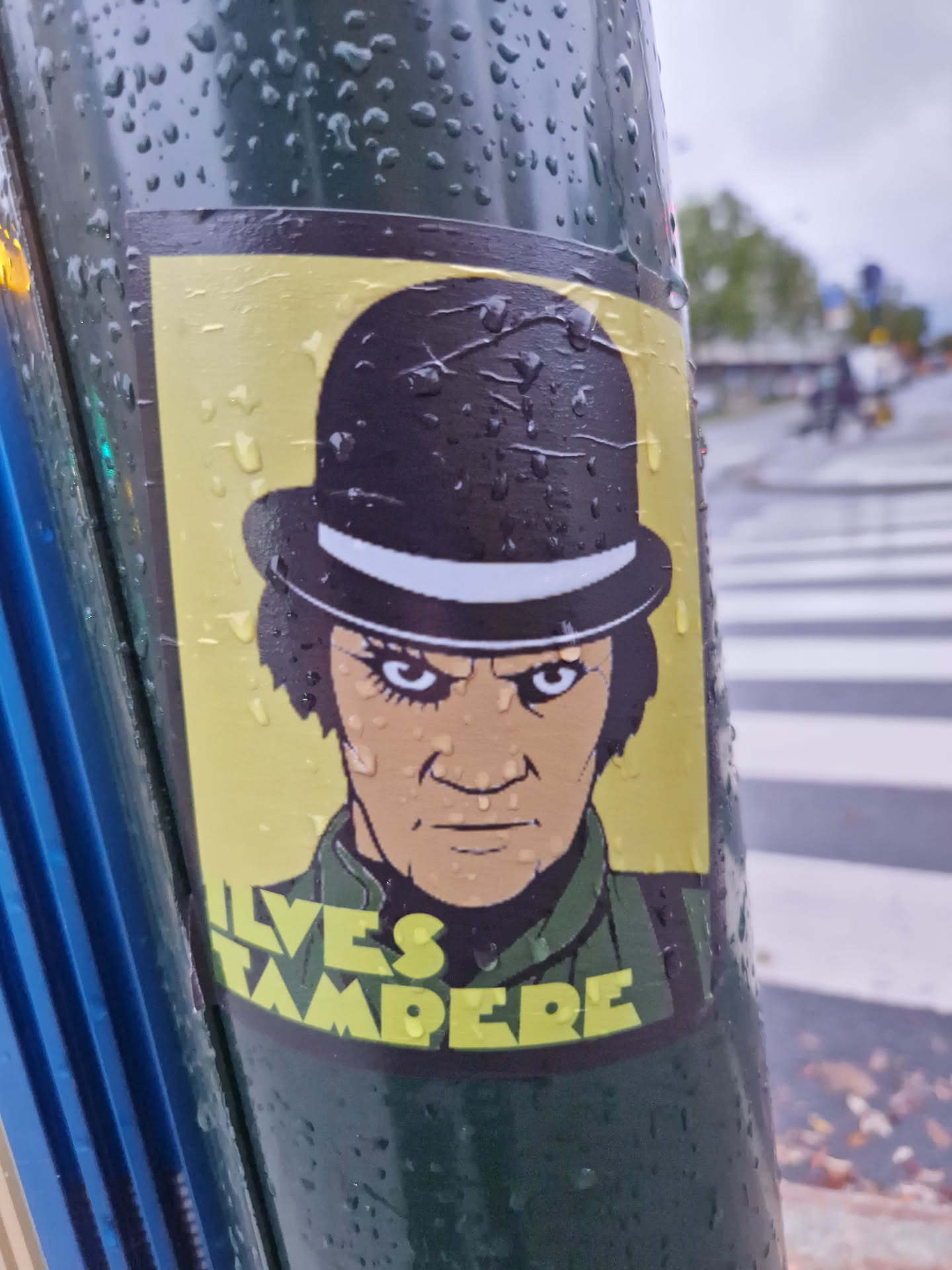 A wet sticker featuring a stylized illustration of a man in a bowler hat, reminiscent of the droogs from Stanley Kubrick's "A Clockwork Orange."  The text "ILVES TAMPERE" is visible at the bottom. The sticker is affixed to a wet, dark green pole.
