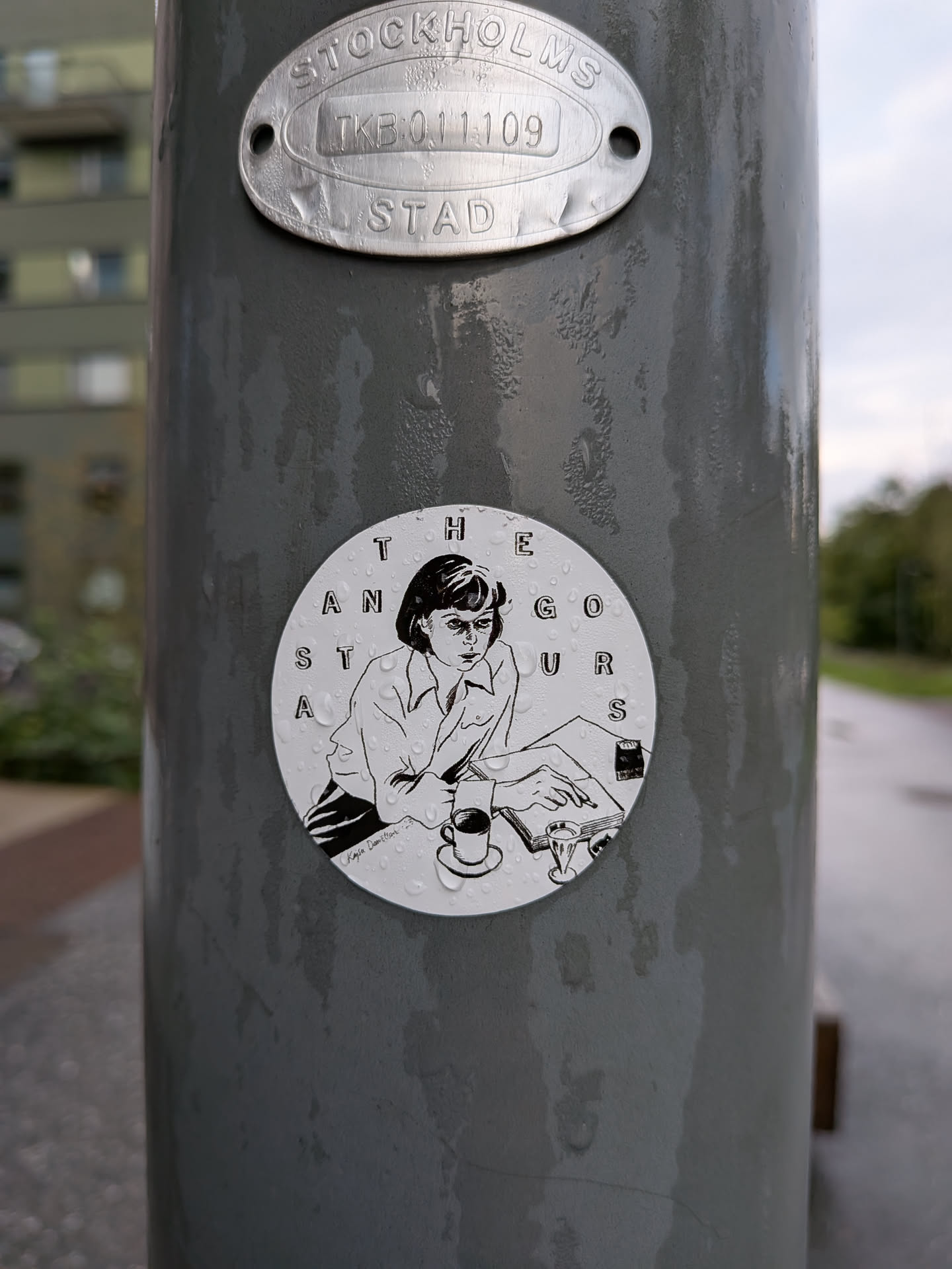 The sticker is circular and features a black and white illustration of a woman sitting at a desk, writing or reading. She has short, dark hair and is wearing a collared shirt. In front of her is an open book, a pen, a cup of coffee on a saucer, and another glass. The text 'THE GORGEOUS ANTS' is arranged in a circle around the illustration. The artist's signature 'Kayla Dameltson' is visible in the lower left corner of the illustration. The sticker is slightly wet, with water droplets visible on its surface.